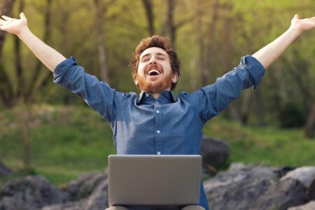 Ein Mann mit Laptop auf - Beruflicher Wechsel dem Schoß im Wald wirft glücklich die Arme hoch und freut sich über neuen Job