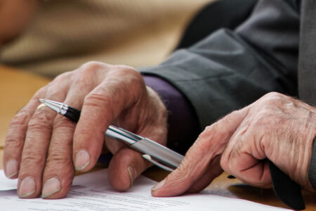 Zwei ältere Hände halten einen Stift und einen Vertrag auf einem Schreibtisch - Erbimmobilien