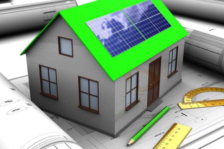 Ein Modell eines Hauses mit grünem Dach und Photovoltaikanlage steht auf einem Bauplan, rundherum liegen weitere Rollen Baupläne, Stifte und Lineale | Energieeffizient bauen
