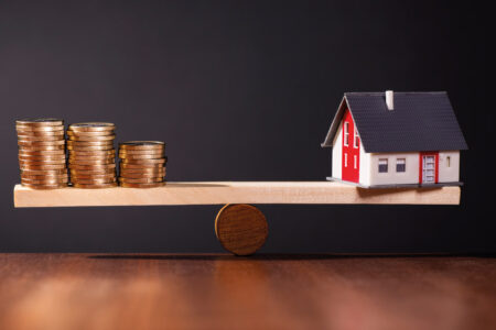 Auf einem Holztisch steht eine Waage aus Holz, die ein Haus gegen Geld abwiegt vor einem dunklen Hintergrund | Immobilienkauf Inflation Zinsen